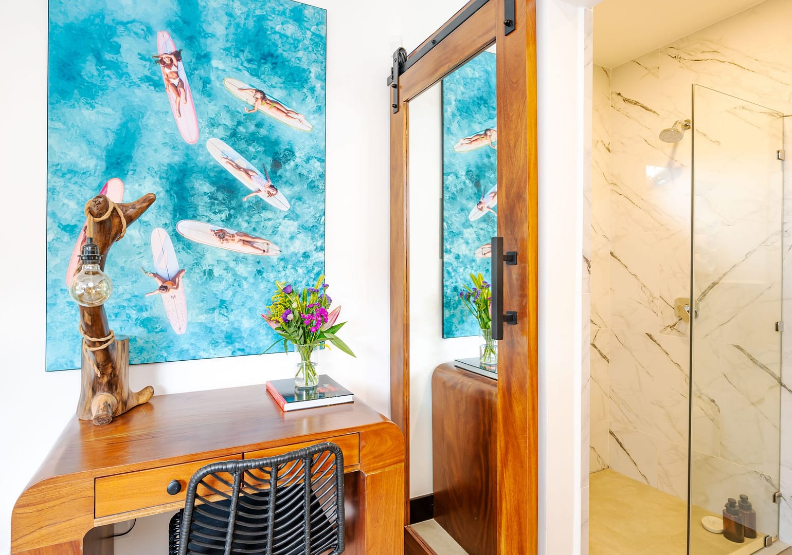 Desk with wall art of a group of female surfers sunbathing on the water atop their boards and sliding barn door with a mirror and view of the ensuite shower